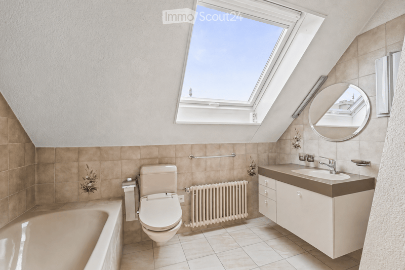 Modern bathroom with a skylight window, beige tiles, a toilet, a sink with a cabinet, a bathtub, and a round mirror.