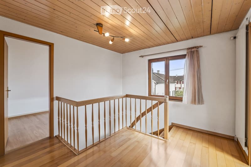 Bright hallway with wooden flooring, white walls, and a wooden staircase leading upwards. Features a window with curtains and recessed ceiling lighting.