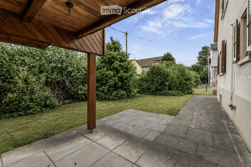 A paved patio area with a wooden pergola on the left, surrounded by green bushes and trees, with a pathway leading to a house on the right.