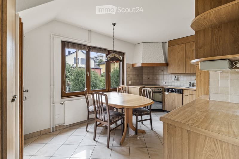 Bright kitchen with wooden cabinets, a dining area with a round table and chairs, and a window offering a view of greenery outside.
