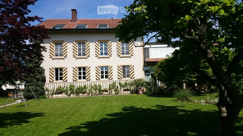 Two-story house with a red-tiled roof, white walls, multiple windows with striped shutters, and a well-maintained garden with a swing set.