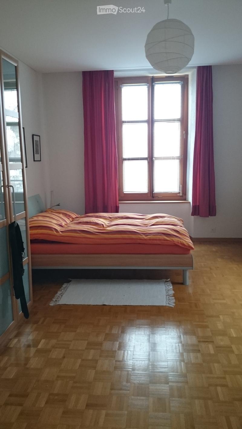 Bright bedroom with wooden flooring, large window with pink curtains, bed with striped bedding, small rug, and a hanging lamp.
