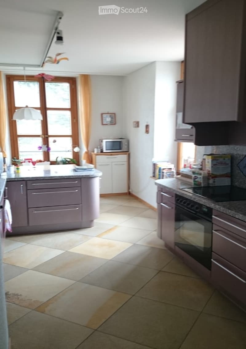 Bright kitchen with white walls, tiled floor, and fitted cabinets. Features a central island with a sink, microwave, and various kitchen items on the counter.