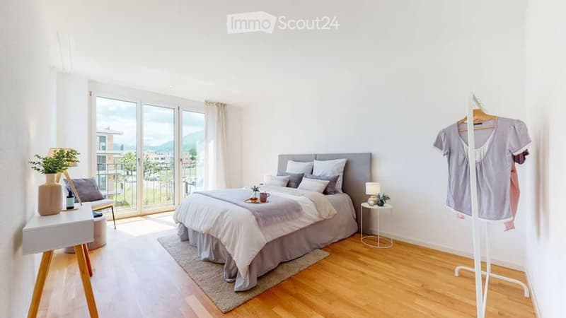 Modern bedroom with a large bed, nightstand, chair, small table, lamp, plant, and a hanger with clothes. Large windows offer a scenic view.