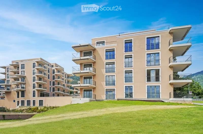 Modern multi-story residential building with balconies and large windows, surrounded by green lawn.