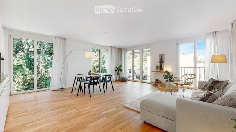 Bright, open-plan living and dining area with wooden flooring, large windows, and sliding glass doors leading to a balcony. Features a white sofa, coffee table, dining table with chairs, floor lamp, and side table with decor.