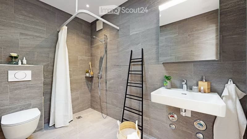 Modern bathroom with grey tiled walls and floor, featuring a toilet, sink with mirror, shower with curtain, and decorative ladder and plant.