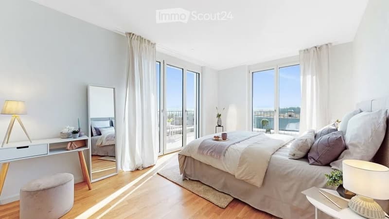 Modern bedroom with large windows and balcony access, featuring a bed, nightstand, lamp, mirror, and a small seating area with a stool and side table.