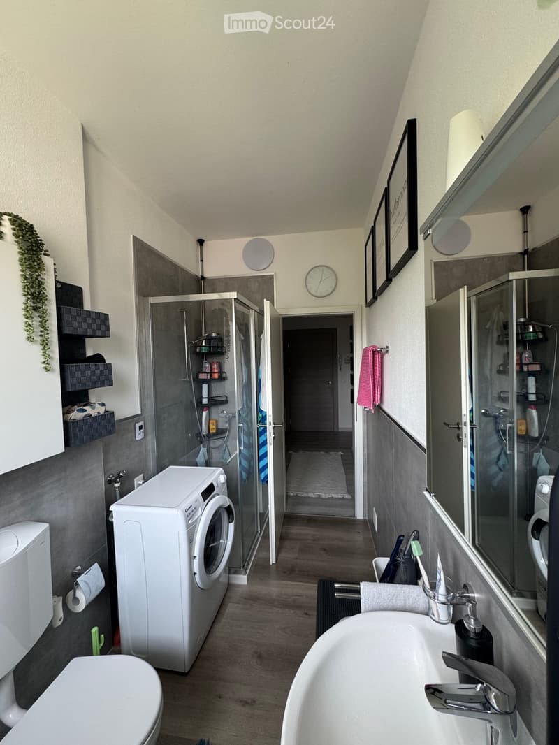 Modern bathroom with a bathtub, toilet, washing machine, shower, and shelves. Features a sleek design with grey tiles and wooden flooring.