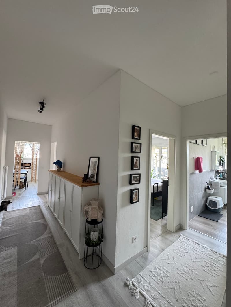 A modern hallway with light grey walls, wooden flooring, and several framed pictures on the wall. There is a small shelf with decorative items, a pet litter box in the adjacent bathroom, and a glimpse of a living area with a table and chairs.
