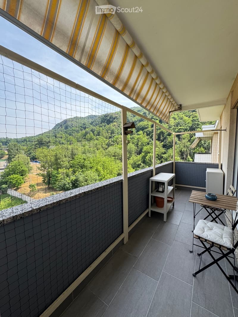 Balcony with tiled floor, small table, chair, shelf, and potted plant, overlooking a green landscape.
