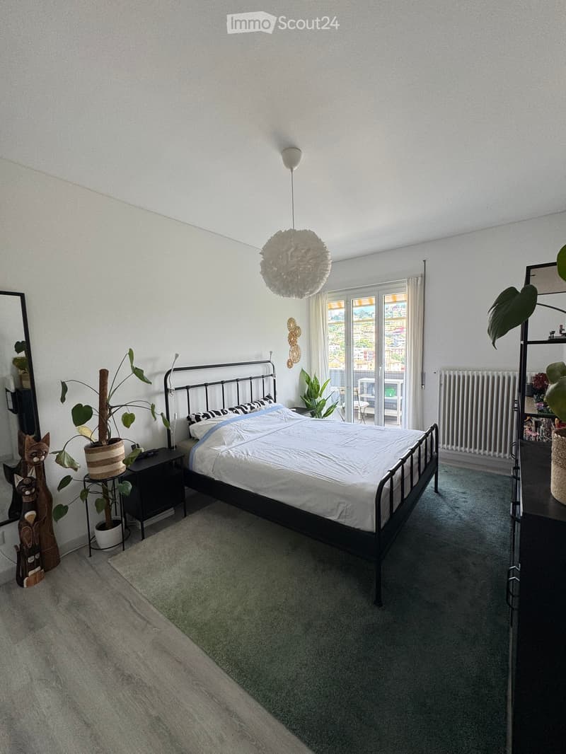 Modern bedroom with a double bed, nightstand, mirror, potted plants, and a hanging lamp. The room features large windows with a view and a radiator.