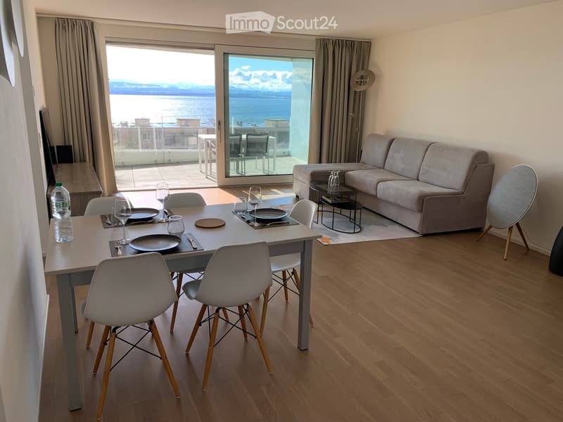 Modern living room with a sofa, coffee table, dining table, chairs, and wine glasses, overlooking a scenic view.