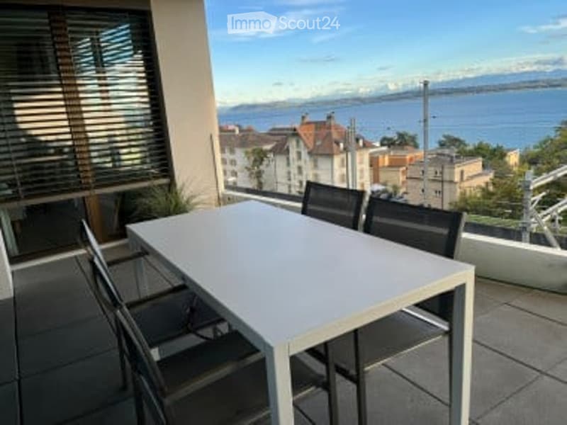 Balcony with a modern white dining table and four black chairs, overlooking a scenic lake and distant mountains.