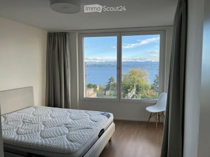 Bright bedroom with large windows offering a scenic view, featuring a bed and a white chair.