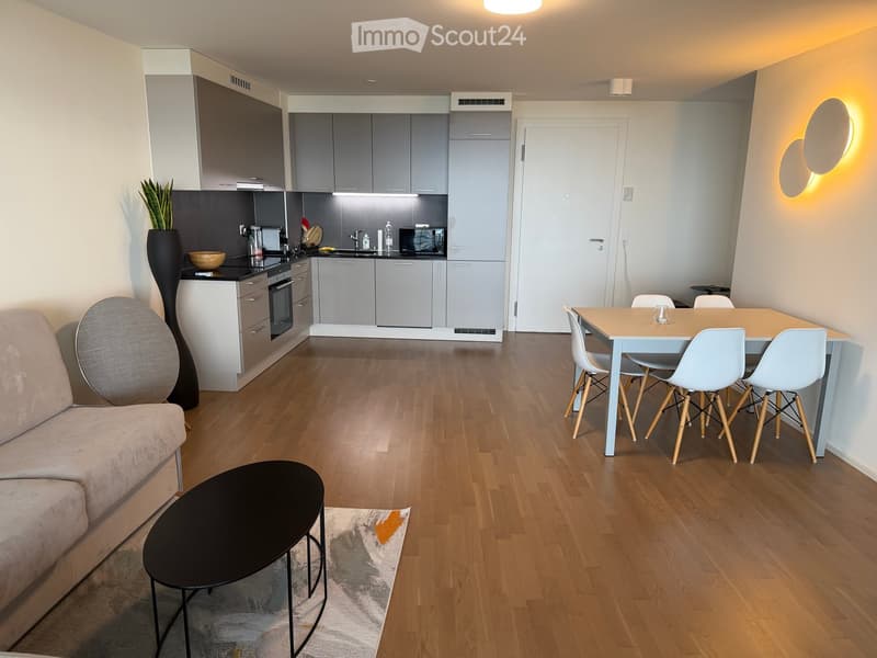 Modern open-plan living and dining area with a sofa, coffee table, dining table with chairs, and a fully equipped kitchen with cabinets, oven, and microwave.