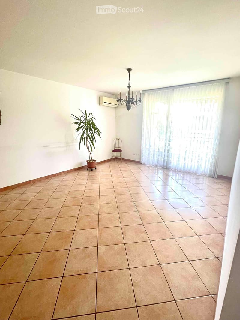 Empty room with tiled floor, white walls, large window with sheer curtains, air conditioning unit, chandelier, small decorative chair, and potted plant.