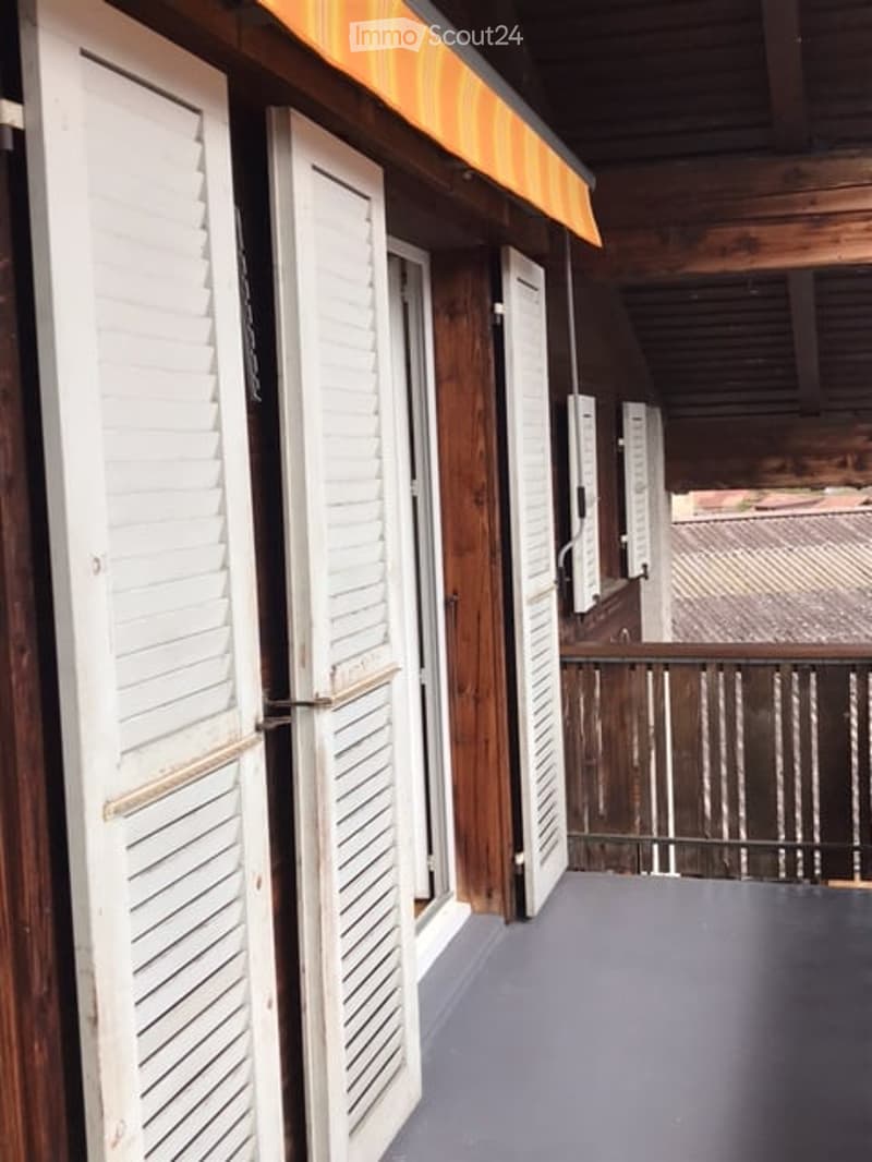 Wooden balcony with white shutters and a metal railing, featuring a grey floor and a wooden ceiling.