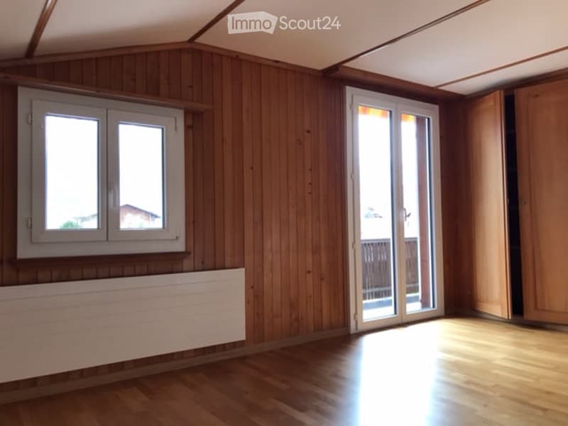 Empty room with wooden walls and floor, large window, sliding glass door leading to a balcony, radiator, and built-in wooden closet.