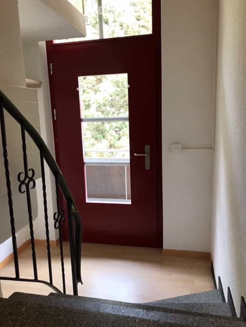 A modern hallway with a wooden floor, white walls, and a staircase with a black wrought-iron railing. A red front door with a glass panel is visible at the end of the hallway.