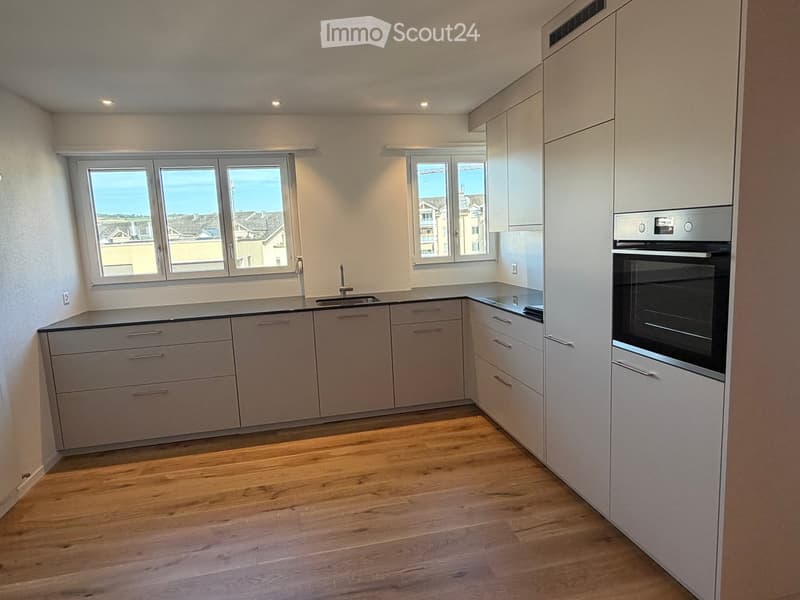 Modern kitchen with light-colored cabinets, wooden flooring, and a stainless steel oven. Features a sink and ample natural light from large windows.