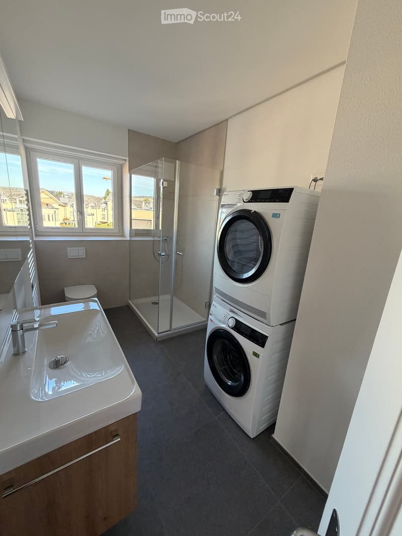 Modern bathroom with a sink, toilet, glass-enclosed shower, and stacked washer/dryer unit. Large windows provide natural light.