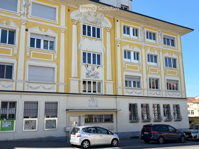 A multi-story building with yellow and white facade, decorative columns, and ornate carvings. Windows have shutters, and there is a lower section with a door and posters.