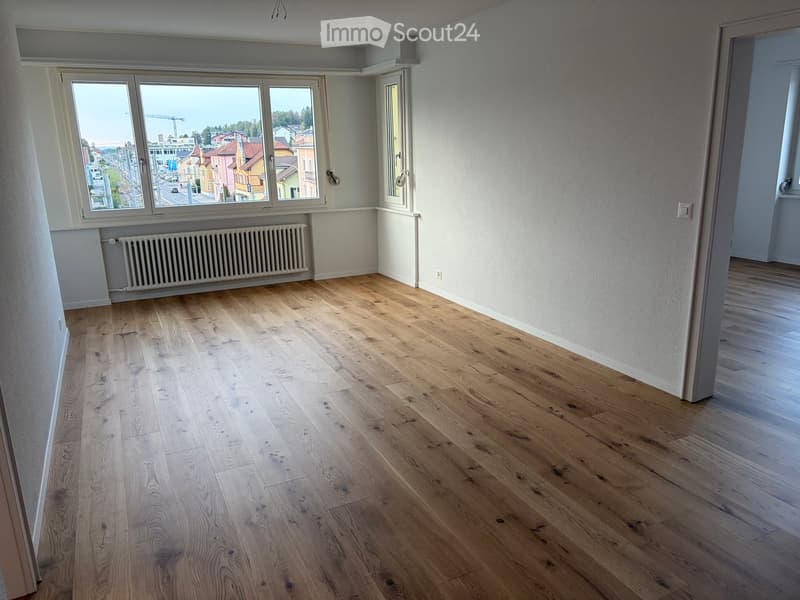 Empty room with light wooden flooring, white walls, large windows offering a view of the city, and a radiator under one window.