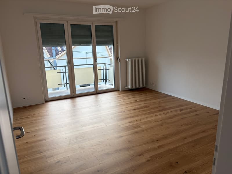 Empty room with wooden flooring, large sliding glass doors leading to a balcony, and a radiator on the wall.