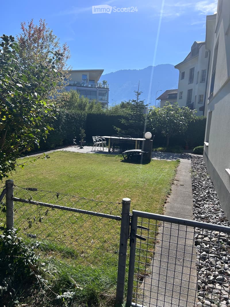 A fenced garden with a grassy lawn, a patio area with a table and chairs, and a view of surrounding buildings and mountains.