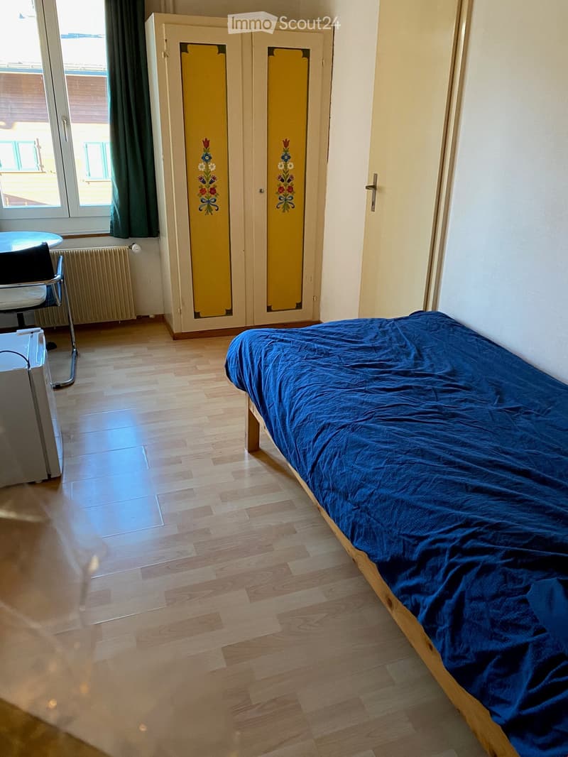 Single bed with blue blanket, yellow floral wardrobe, round table, chair, white radiator, glass window with green curtains