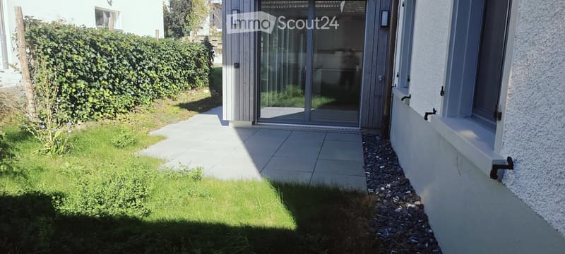 Small garden area with a tiled pathway leading to a glass sliding door of a house, surrounded by a hedge and grass.