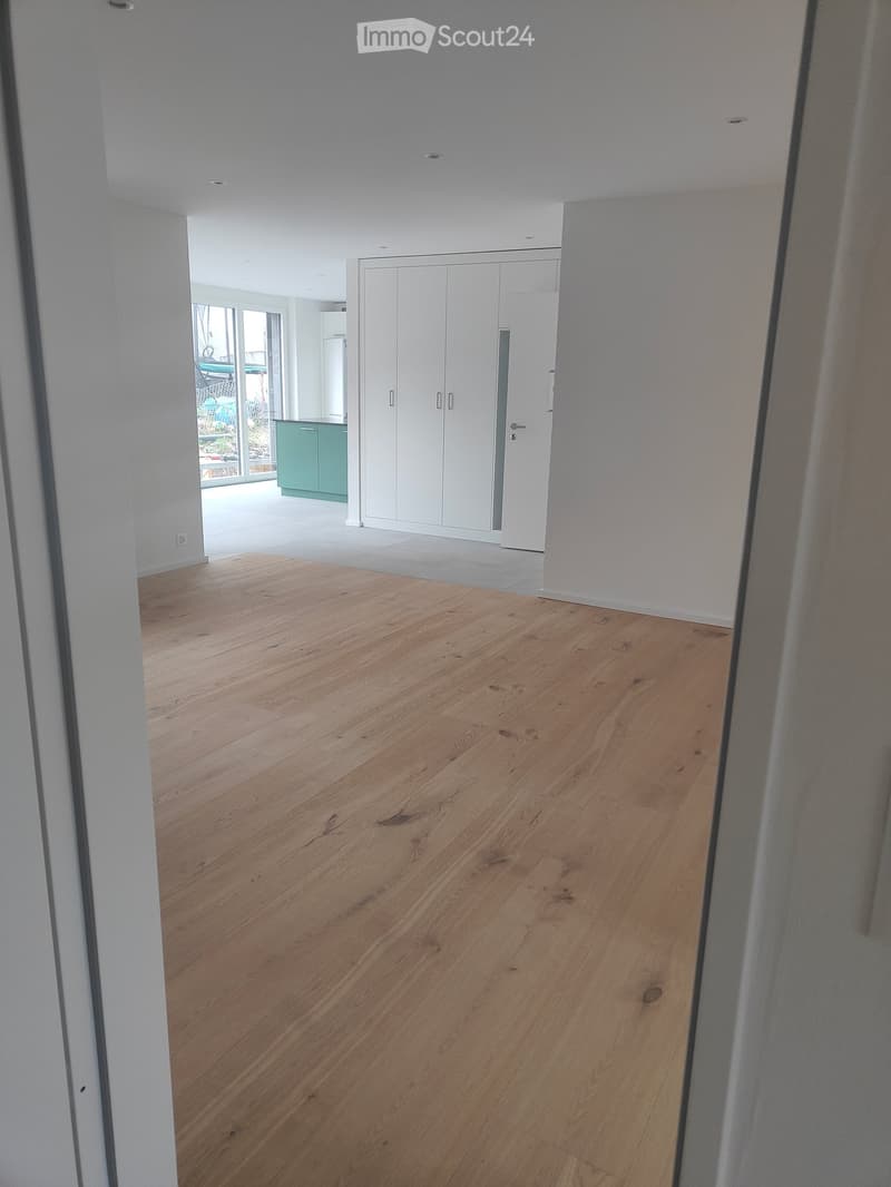 The image shows an empty room with wooden floors, white walls, and a white door leading to a glass wall on the left side. The room also has a white built-in wardrobe and a power outlet on the left side.