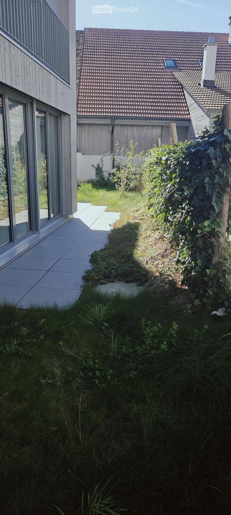 Garden area with a raised ground floor, featuring a pathway, grass, and plants, adjacent to a modern house with large windows and a tiled roof.