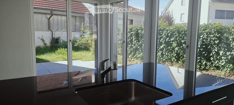 Modern kitchen with large windows overlooking a garden, featuring a stainless steel sink and faucet.