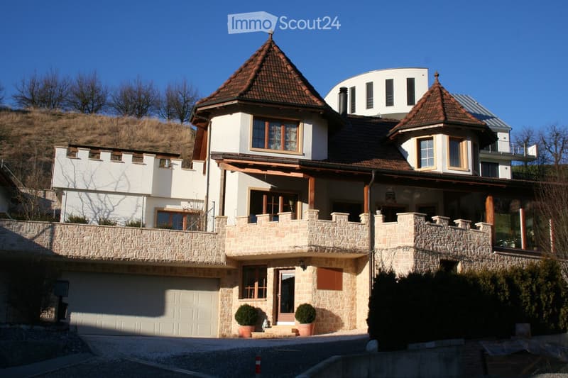 A multi-story house with a stone facade, wooden accents, and a tiled roof. Features include a garage, balcony, and surrounding greenery.