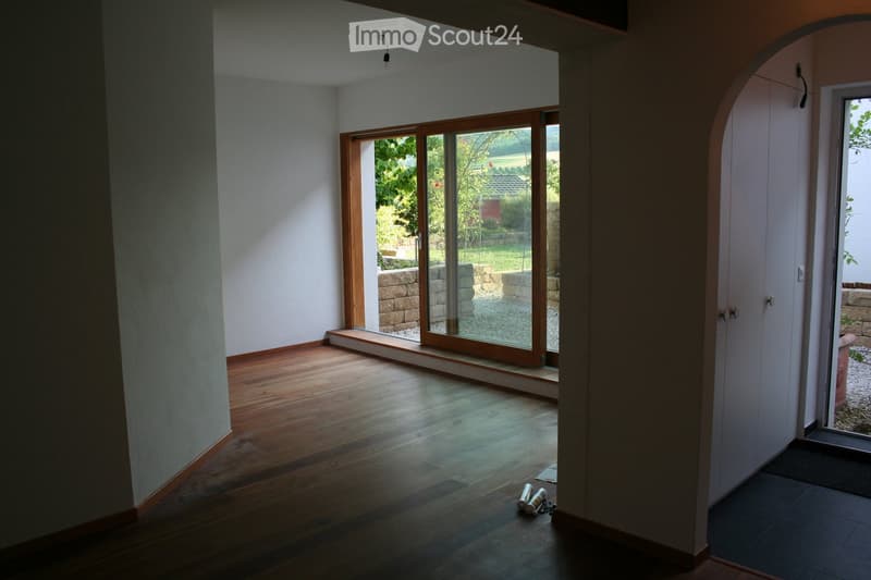 Empty room with wooden flooring, large sliding glass doors leading to a garden, and a white wall with an arched doorway.