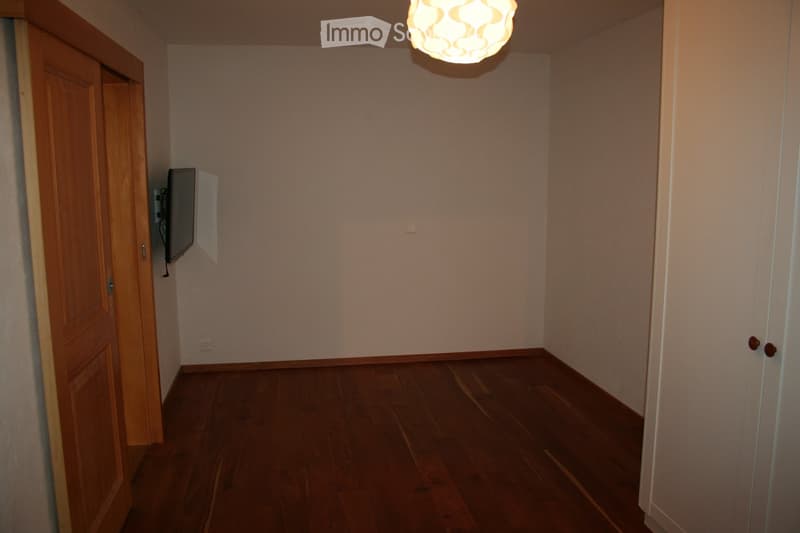 Empty room with wooden flooring, white walls, and a ceiling light fixture. A flat-screen TV is mounted on one wall, and there are two doors, one on each side.