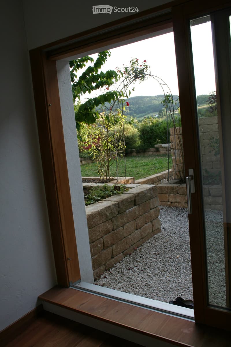 Open wooden-framed window or balcony door showing a garden with stone walls, gravel, and plants.