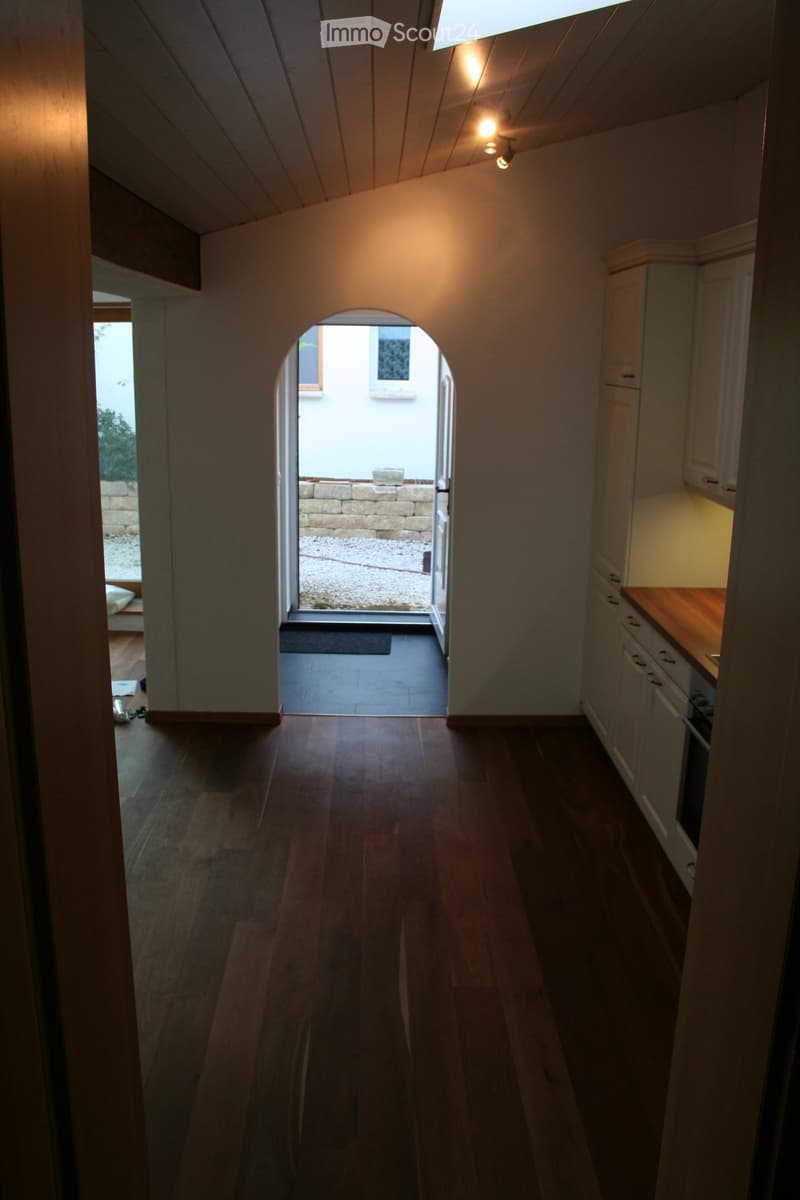 A narrow hallway with wooden flooring, white walls, and a skylight ceiling. An arched doorway leads to an outdoor patio area with a stone wall and gravel flooring.