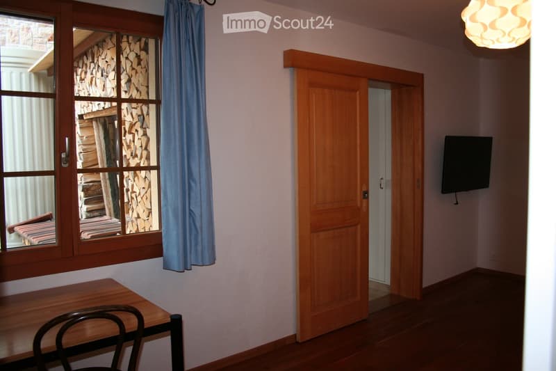 A small, unfurnished room with a wooden table, a chair, and a TV mounted on the wall. The room has a large window with a view of stacked firewood and a blue curtain.