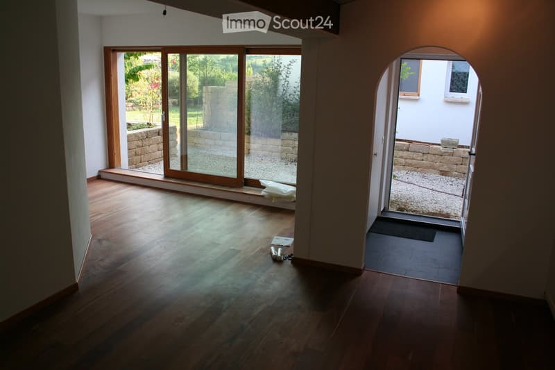 Empty room with wooden flooring, large sliding glass doors leading to a garden area, and a small entryway with a tiled floor.