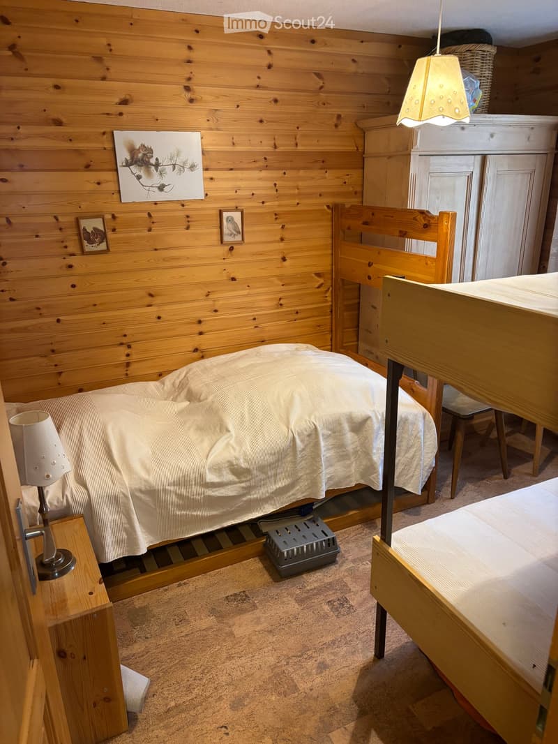 Cozy bedroom with wooden walls, two single beds, a small table, a chair, and a lamp. The room features a pet carrier and decorative artwork on the wall.