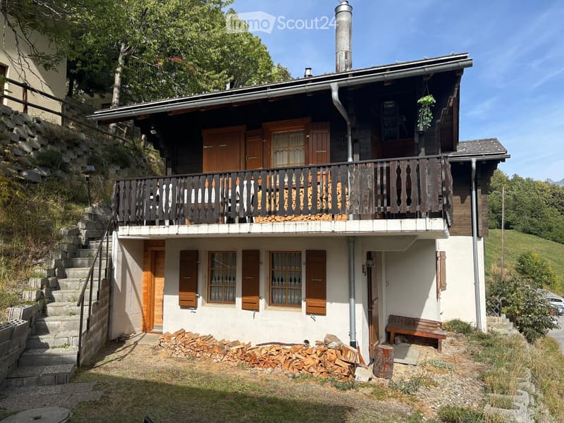 Two-story wooden house with a balcony, stone steps, and a stack of firewood in front.