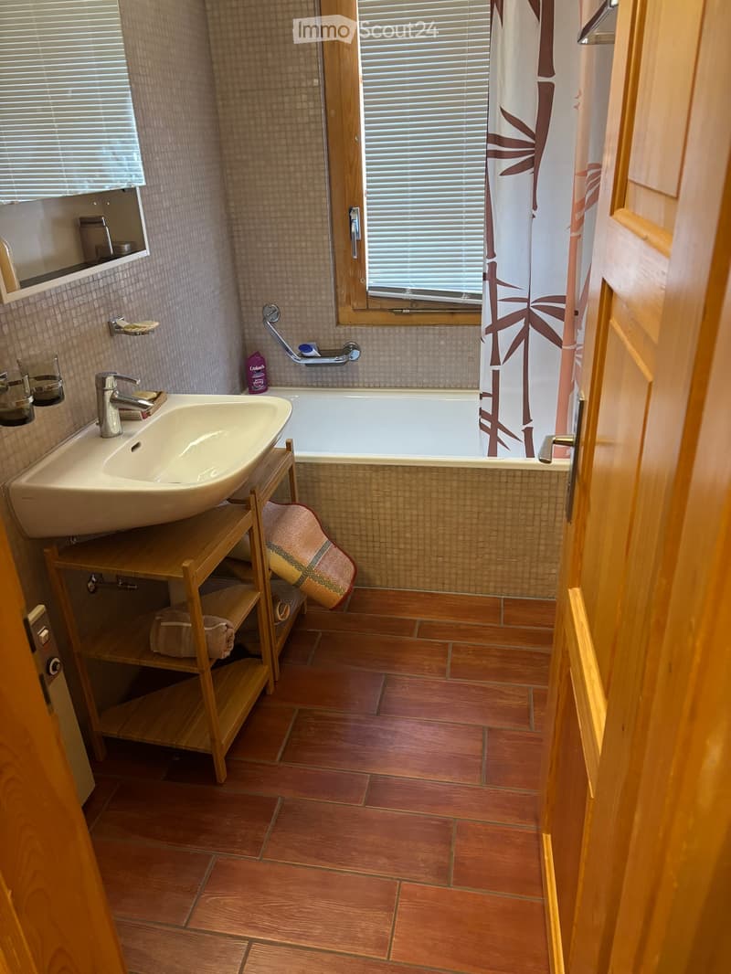 Bathroom with a white sink, bathtub, wooden shelf, and a shower curtain with a bamboo design. The floor is tiled in a warm, reddish-brown color, and the walls are tiled with small, light-colored tiles.