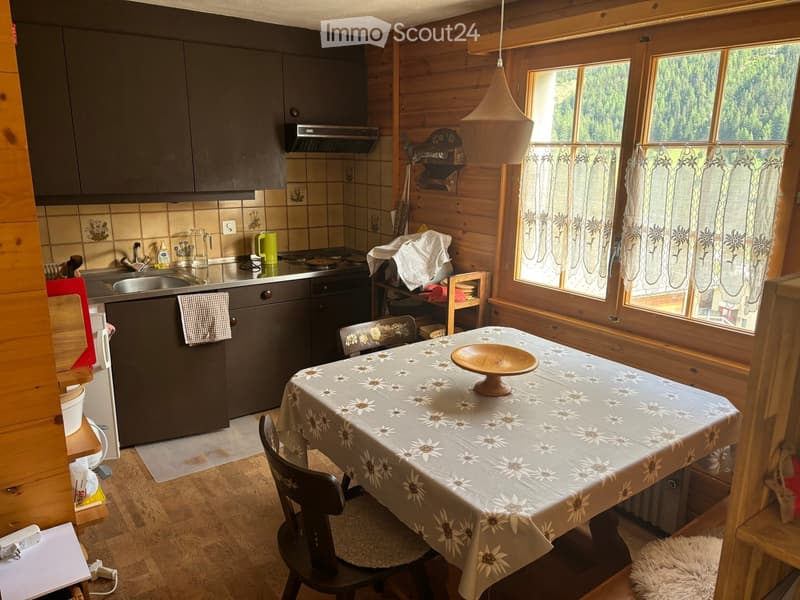 A cozy kitchen with wooden walls, a dining table with a floral tablecloth, and modern appliances including a sink, refrigerator, and oven. The room features large windows with floral curtains and a view of greenery outside.