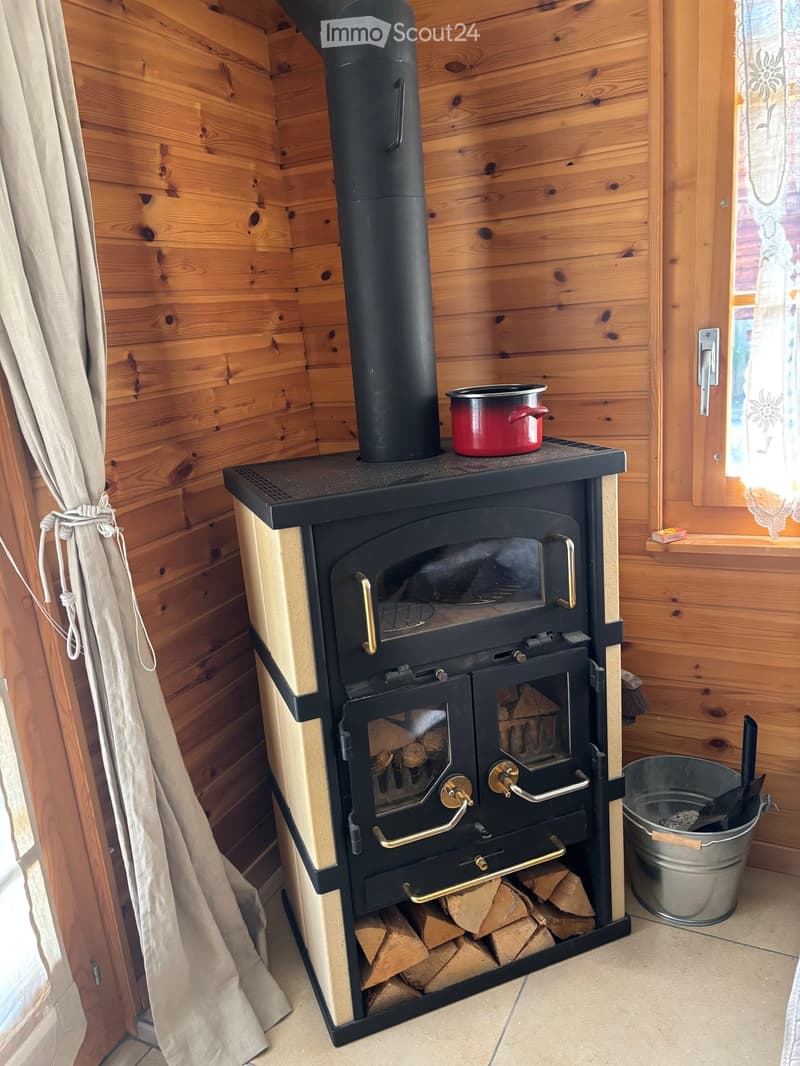 Wooden-paneled room with a black wood-burning stove, a red pot on top, and a stack of firewood underneath.