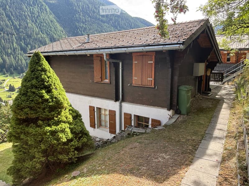A two-story wooden house with brown shutters and a tiled roof, situated in a mountainous area with a well-maintained garden and a view of the surrounding landscape.