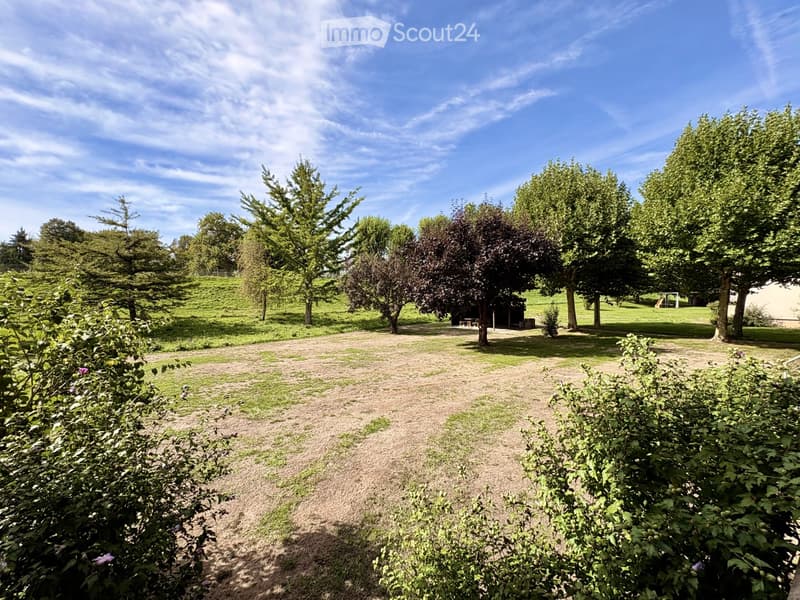 A spacious garden area with a gravel pathway, surrounded by trees and shrubs, with a small shed and a goalpost visible in the background.