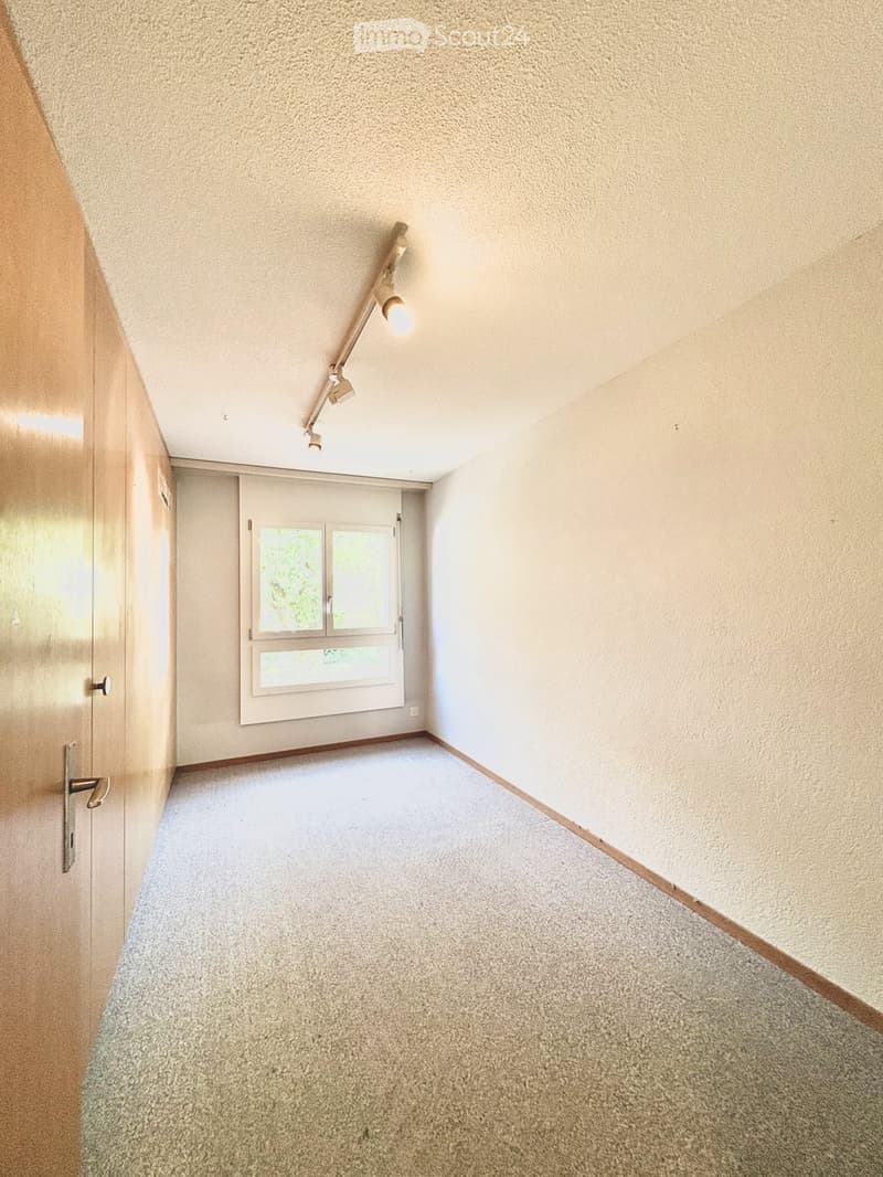 Empty room with beige walls, carpeted floor, and a single window with blinds. Features a wooden door and a track light fixture on the ceiling.
